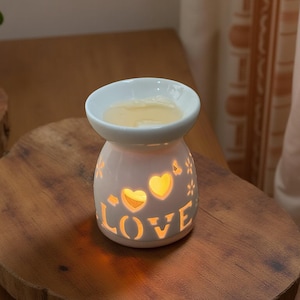May include: A white ceramic essential oil diffuser with the word "LOVE" cut out, illuminating the word and heart shapes. The diffuser sits on a wooden surface. The top bowl contains a yellow liquid.