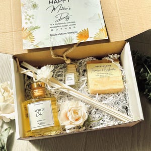 Könnte beinhalten: Eine Geschenkbox zum Muttertag mit einem Schilfrohrdiffusor, Seife und Duft. Die Box enthält eine dekorative Blume und eine Karte mit der Aufschrift "Happy Mother's Day". Der Schilfrohrdiffusor ist mit "White Cafè" beschriftet. Die Seife ist mit "Turmeric & Cashmere" beschriftet.