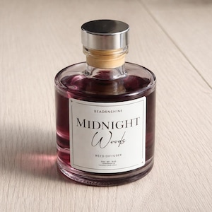 Op de afbeelding: Een heldere glazen rietverspreiderfles gevuld met een dieppaarse vloeistof. De fles heeft een zilveren dop en een etiket met de tekst "Midnight Woods" en "Reed Diffuser". Het product is van "Beadsnshine".