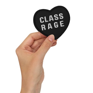 May include: A heart-shaped black embroidered patch with a thick black border. The text CLASS RAGE is stitched in white capital letters across the center of the patch. The design features a clean, bold aesthetic with high-contrast white lettering.