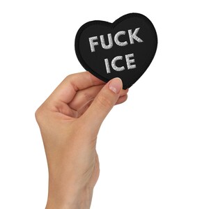 May include: A heart-shaped black embroidered patch with a thick black border. The text FUCK ICE is stitched in white capital letters across the centre of the patch. The design features a clean, bold aesthetic with high-contrast white lettering.