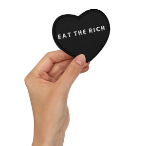 Puede incluir: Un parche negro en forma de corazón con el texto bordado "EAT THE RICH" en blanco. El parche está sostenido por una mano sobre un fondo blanco. El parche es probablemente de tela.