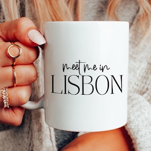 May include: White ceramic mug with the text "meet me in LISBON" in black script. The mug is held by a person wearing a gray sweater and multiple rings. The mug is a standard size for coffee or tea.