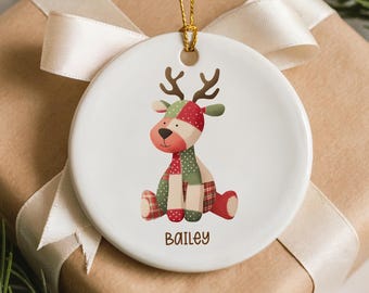 Puede incluir: Adorno navideño de cerámica blanca con un reno de dibujos animados con un diseño de patchwork en rojo, verde y marrón. El reno está sentado sobre un fondo blanco, con el nombre "Bailey" impreso debajo. El adorno cuelga de un hilo dorado, encima de un regalo envuelto.