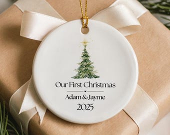 May include: A white ceramic Christmas ornament with a gold string, featuring a watercolor Christmas tree design. The text "Our First Christmas" is above the names "Adam & Jayme" and the year "2025". The ornament is on a wrapped gift.