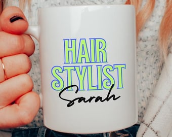 Może przedstawiać: Biały ceramiczny kubek z napisem "HAIR STYLIST Sarah" w niebieskich i zielonych literach. Kubek trzyma osoba nosząca złoty naszyjnik i pierścionek. Kubek jest w standardowym rozmiarze.