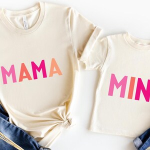 May include: Two cream-colored t-shirts, one adult and one child size, with the words "MAMA" and "MINI" in pink and orange block letters. Blue jeans are visible on either side of the shirts.