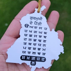 May include: A white pendant in the shape of a map with text in a foreign script. The pendant is held in a hand against a blurred green background. A gold-colored string is attached to the top of the pendant.