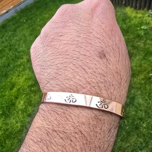 Puede incluir: Una pulsera de color cobre con el símbolo Om grabado. La pulsera tiene un diseño de banda simple, y el símbolo Om se repite en la parte delantera. La pulsera se lleva en la muñeca.
