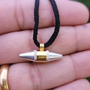 Könnte beinhalten: Eine silber-goldene Anhänger-Halskette mit einer schwarzen geflochtenen Kordel. Der Anhänger hat eine zylindrische Form mit silbernen, spitz zulaufenden Enden und einem goldenen Mittelteil. Die Halskette wird vor einem verschwommenen Hintergrund präsentiert.