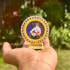 Könnte beinhalten: Ein dekoratives religiöses Objekt mit einem Porträt eines Mannes mit Turban. Das Porträt ist in einem kreisförmigen Rahmen mit roten und klaren Edelsteinen eingefasst. Der Artikel hat einen goldfarbenen Sockel und wird in einer Hand vor einem verschwommenen grünen Hintergrund gehalten.