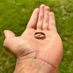 Può includere: Un anello color bronzo con una superficie strutturata. L'anello è tenuto nel palmo di una mano aperta su uno sfondo verde sfocato. Il motivo dettagliato e la forma circolare dell'anello sono chiaramente visibili.