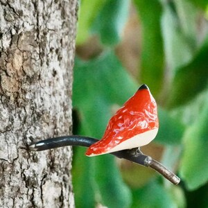 Puede incluir: Tres esculturas de pájaros de cerámica posadas en un tronco de árbol. Los pájaros son blancos, rojos y amarillos, con ramas de metal negro. La corteza del árbol es texturizada y el follaje verde es visible en el fondo. Arte decorativo de jardín.
