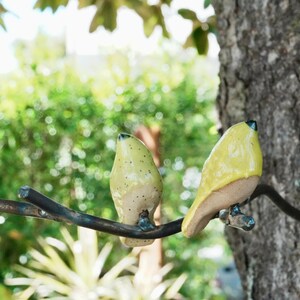 Puede incluir: Dos esculturas de pájaros de cerámica posadas en una rama de metal, montadas en un tronco de árbol. Un pájaro es de color verde claro con manchas oscuras, el otro es amarillo. El fondo es follaje verde borroso.