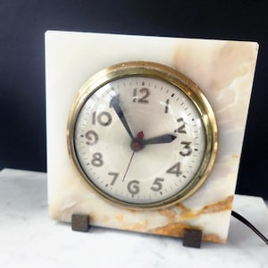 May include: A vintage clock with a gold-tone bezel and a cream-colored face. The clock is set in a rectangular marble base with brown accents and supported by two small, dark-colored feet. The clock hands are black and red.