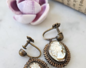 May include: A pair of vintage cameo earrings with a gold tone finish. The earrings feature a white cameo with a woman's profile and are surrounded by a delicate wire design.