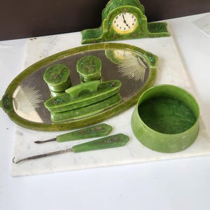 May include: A collection of vintage green vanity items, including a clock, a mirrored tray with containers, and grooming tools. The items are arranged on a white marble surface, showcasing a cohesive set.