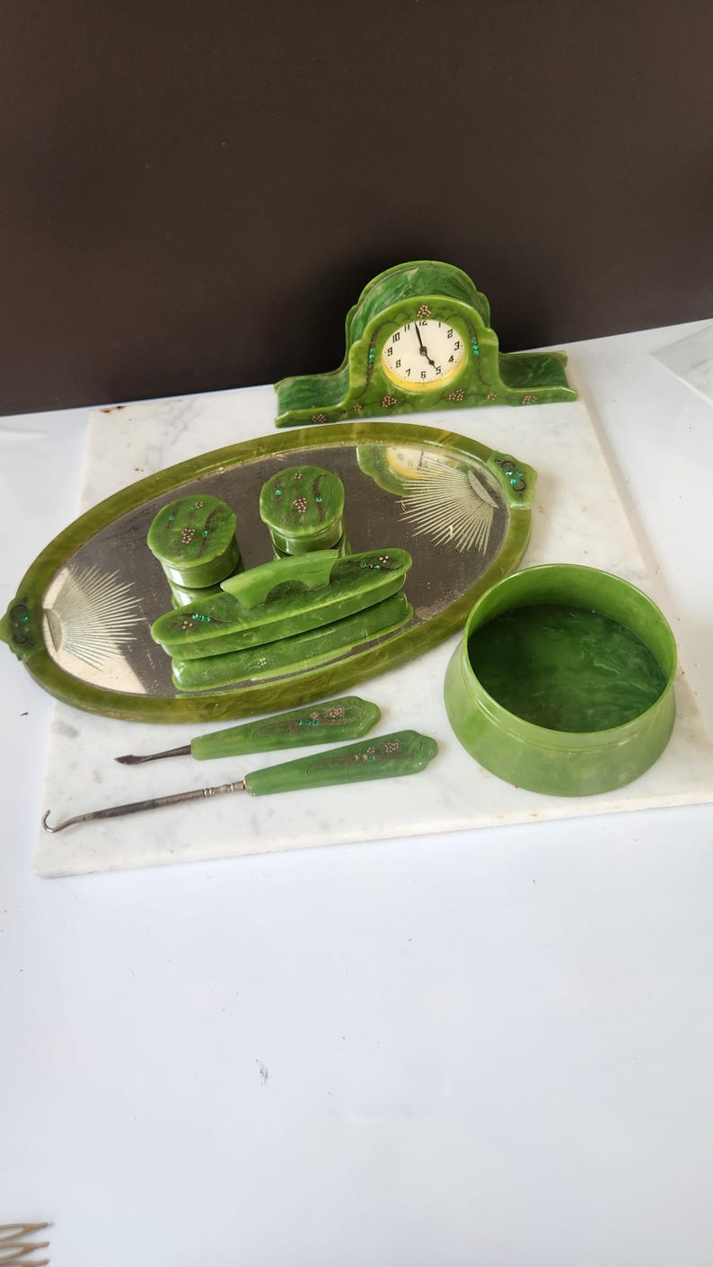 May include: A collection of vintage green vanity items, including a clock, a mirrored tray with containers, and grooming tools. The items are arranged on a white marble surface, showcasing a cohesive set.