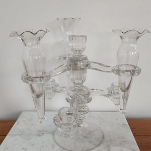 May include: A clear glass candelabra with multiple arms and candle holders. The candelabra has a central stem with a circular base and several arms extending outwards, each holding a candle cup. The design features a delicate, ornate style.