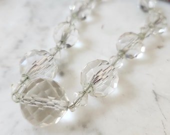 May include: A clear, faceted crystal bead necklace. The beads are strung together with a thin, silver-colored wire. The necklace is displayed on a white marble surface. The beads are round and reflect light, creating a sparkling effect.