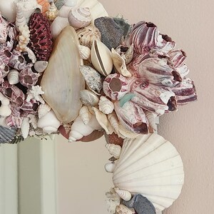 May include: A decorative mirror frame adorned with a variety of seashells in shades of white, cream, purple, and gray. The frame is densely packed with different types of shells, creating a textured and coastal aesthetic. The mirror is ideal for a beach-themed decor.