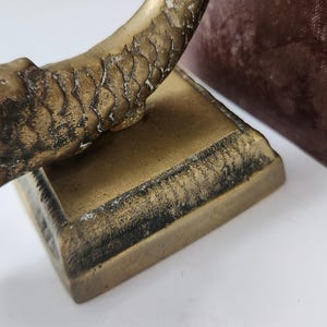 May include: Close-up of a vintage brass sculpture with a textured, scale-like pattern on a curved element. The sculpture sits on a square base with a dark, aged patina. The background includes a glimpse of a dark, textured surface.
