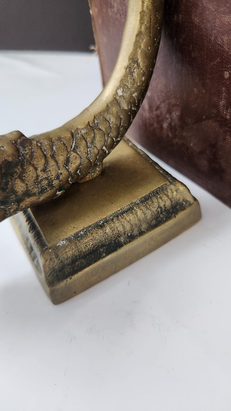 May include: Close-up of a vintage brass sculpture with a textured, scale-like pattern on a curved element. The sculpture sits on a square base with a dark, aged patina. The background includes a glimpse of a dark, textured surface.