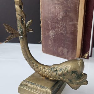 May include: A vintage brass bookend in the shape of a dolphin. The bookend has a square base and a curved body with a fish-like head and tail. Two antique books are in the background.