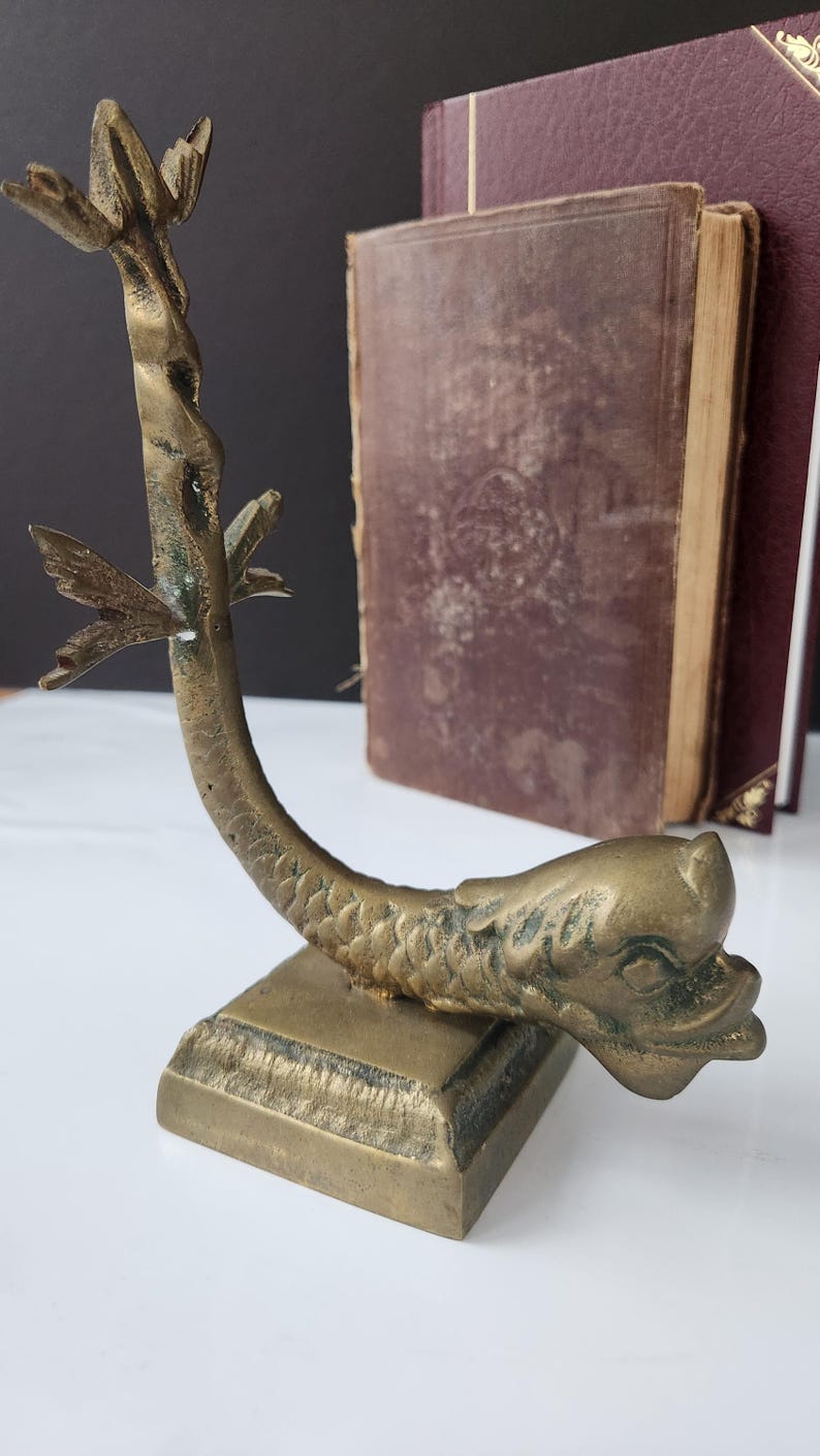 May include: A vintage brass bookend in the shape of a dolphin. The bookend has a square base and a curved body with a fish-like head and tail. Two antique books are in the background.