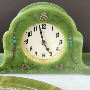May include: Green Art Deco clock with a white clock face and black numerals. The clock has decorative accents, including small green jewels and floral designs. The clock is made of a marbled green material.