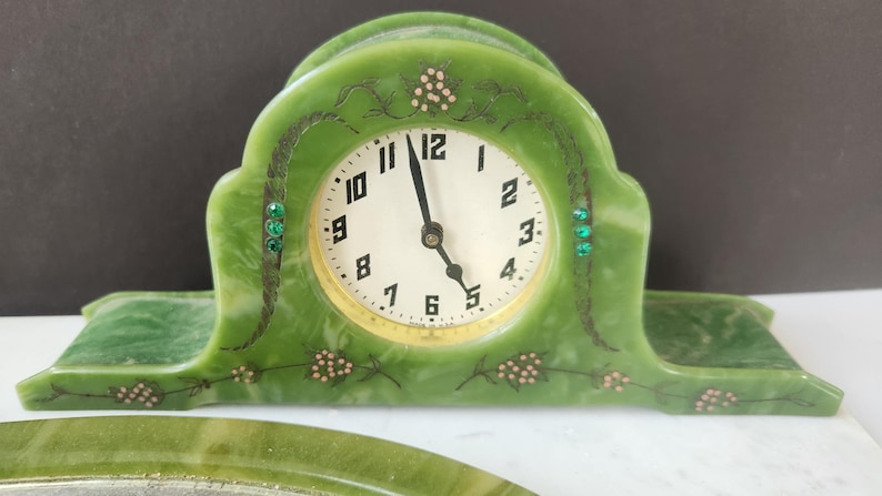 May include: Green Art Deco clock with a white clock face and black numerals. The clock has decorative accents, including small green jewels and floral designs. The clock is made of a marbled green material.
