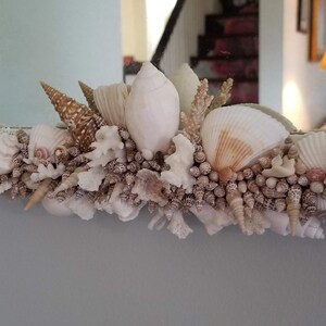May include: A mirror frame decorated with a variety of seashells, coral, and small, round shells. The shells are in shades of white, cream, and brown, creating a coastal or nautical theme. The frame is likely made of wood or a similar material.