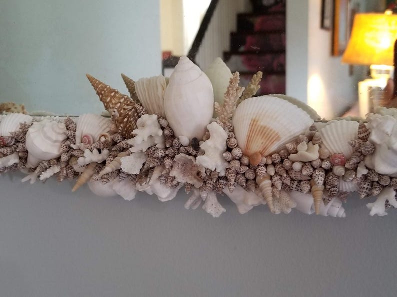 May include: A mirror frame decorated with a variety of seashells, coral, and small, round shells. The shells are in shades of white, cream, and brown, creating a coastal or nautical theme. The frame is likely made of wood or a similar material.