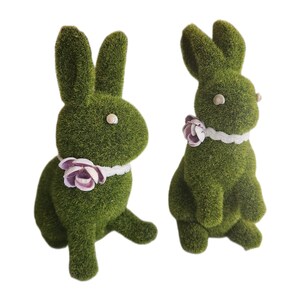 May include: Two green bunny ornaments with a textured, grassy appearance. Each bunny has a white collar with a pink and purple flower. The bunnies have white eyes and are posed in a sitting position.
