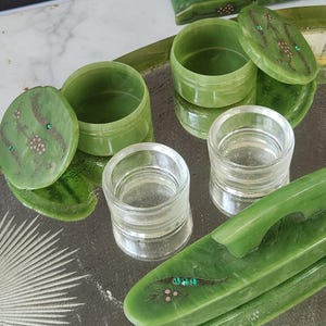 May include: A collection of green jadeite vanity items, including small lidded jars, clear glass containers, and decorative pieces, arranged on a mirrored tray. The jadeite pieces feature floral and gem accents.