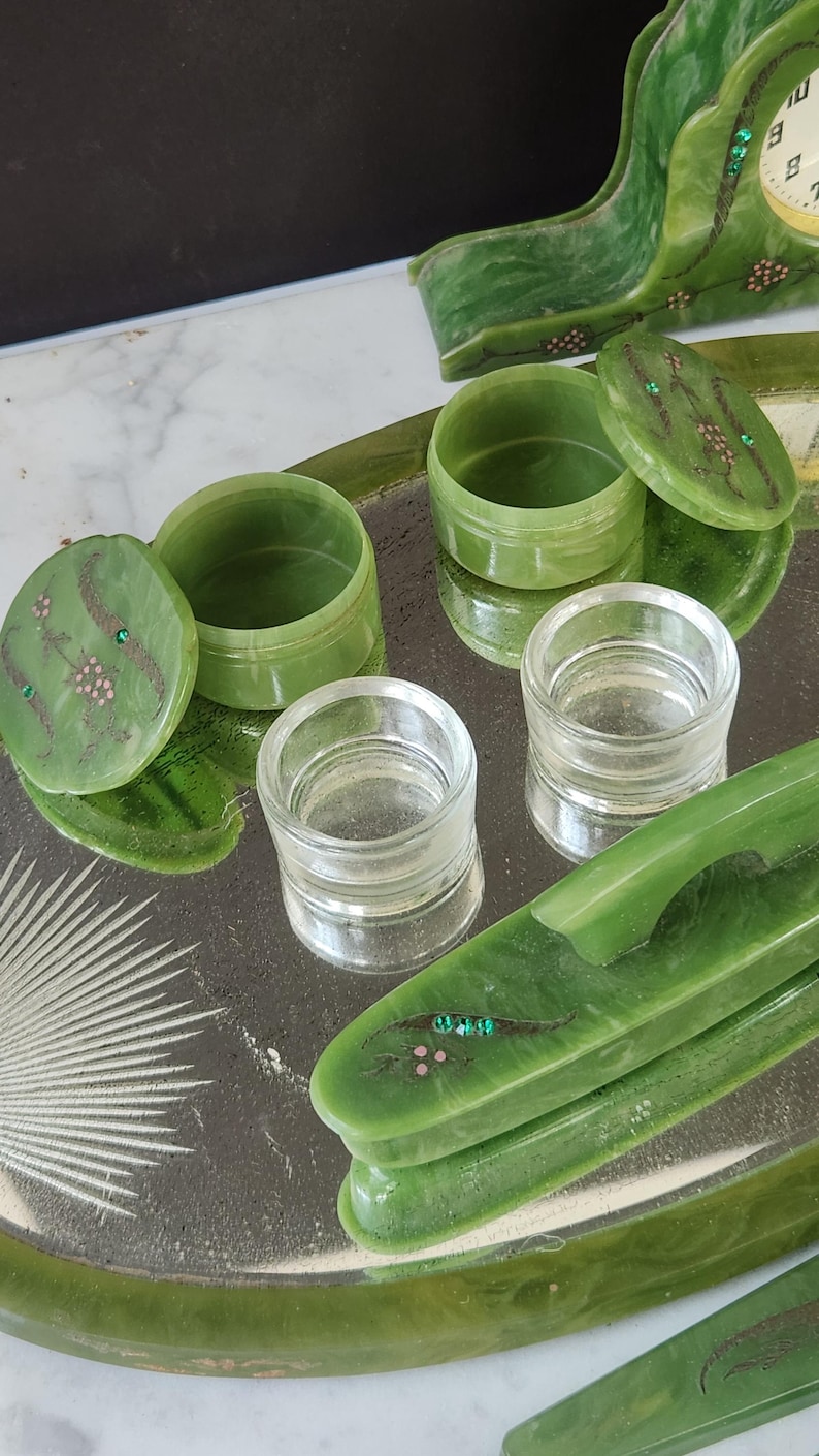 May include: A collection of green jadeite vanity items, including small lidded jars, clear glass containers, and decorative pieces, arranged on a mirrored tray. The jadeite pieces feature floral and gem accents.