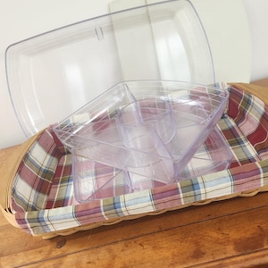 May include: A wicker picnic basket with a plaid lining, holding clear plastic serving trays and a lid. The basket is on a wooden surface, suitable for outdoor dining or food storage.