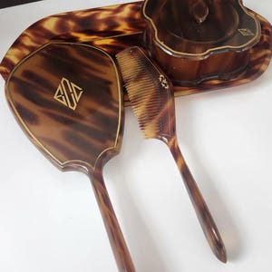 May include: A vintage vanity set with a tortoiseshell pattern. The set includes a hand mirror, comb, a lidded jar, and a tray. The mirror and comb have gold-colored accents and the mirror has a gold monogram. The set is on a white surface.