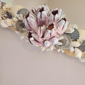 May include: A decorative mirror frame adorned with a variety of seashells, coral, and a large, textured, barnacle-like cluster in shades of pink and white. The frame features a natural, coastal theme with a mix of textures and colors.
