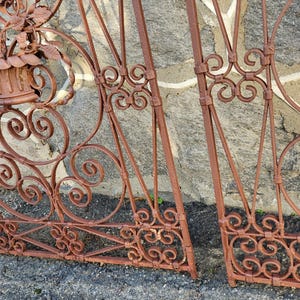 Pu&ograve; includere: Sezioni di cancello in ferro battuto marrone ruggine con decorazioni a volute e accenti floreali. Il cancello presenta un aspetto usurato, che suggerisce l'uso all'aperto. Una piccola pala verde &egrave; visibile sullo sfondo.