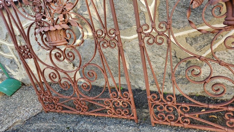 Pu&ograve; includere: Sezioni di cancello in ferro battuto marrone ruggine con decorazioni a volute e accenti floreali. Il cancello presenta un aspetto usurato, che suggerisce l'uso all'aperto. Una piccola pala verde &egrave; visibile sullo sfondo.