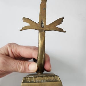 May include: A vintage brass cross with a stylized design. The cross has a rectangular base and a vertical stem with horizontal and upward-pointing elements. The brass has a warm, aged patina, suggesting its antique nature.