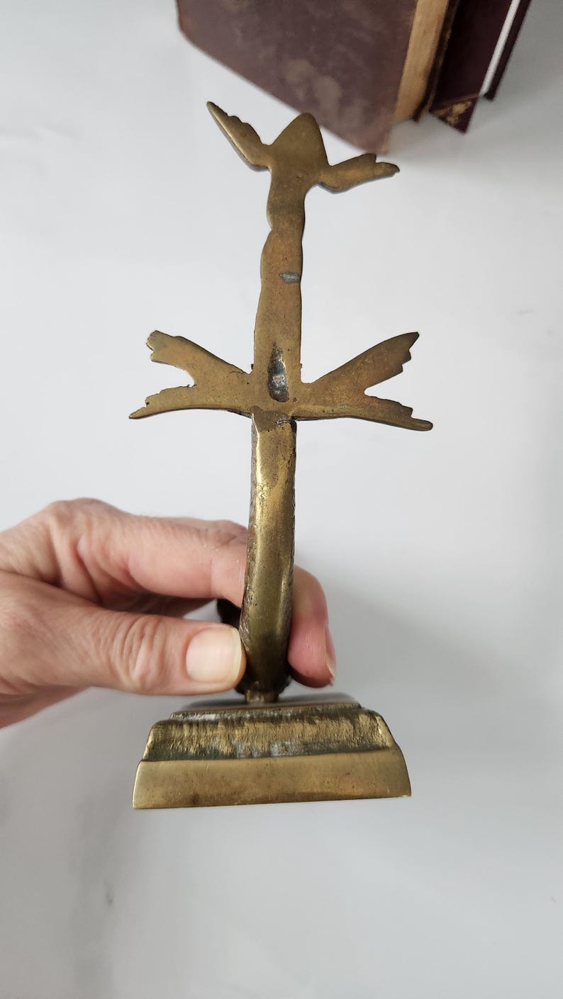 May include: A vintage brass cross with a stylized design. The cross has a rectangular base and a vertical stem with horizontal and upward-pointing elements. The brass has a warm, aged patina, suggesting its antique nature.