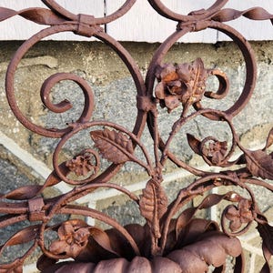 Pu&ograve; includere: Pannello decorativo in ferro battuto marrone arrugginito con motivi floreali e a volute. Il design include rose stilizzate, foglie e un elemento centrale a forma di cestino. La lavorazione del metallo ha un aspetto invecchiato, suggerendo l'uso all'aperto.