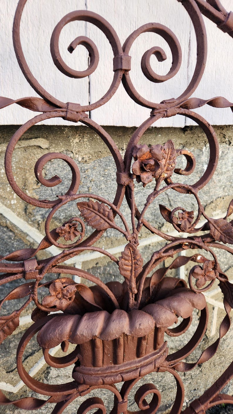Pu&ograve; includere: Pannello decorativo in ferro battuto marrone arrugginito con motivi floreali e a volute. Il design include rose stilizzate, foglie e un elemento centrale a forma di cestino. La lavorazione del metallo ha un aspetto invecchiato, suggerendo l'uso all'aperto.