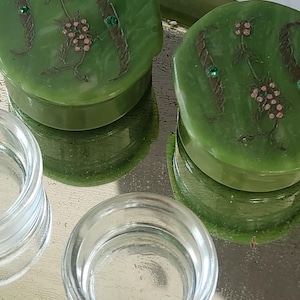May include: Two green, round trinket boxes with floral designs and small green gems. The boxes sit on a reflective surface next to two clear glass containers. The boxes have a raised edge and a smooth, glossy finish.