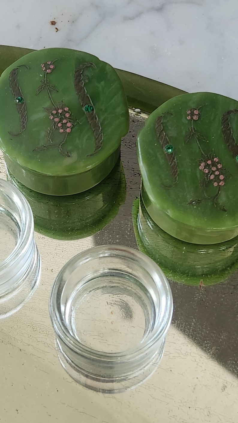 May include: Two green, round trinket boxes with floral designs and small green gems. The boxes sit on a reflective surface next to two clear glass containers. The boxes have a raised edge and a smooth, glossy finish.