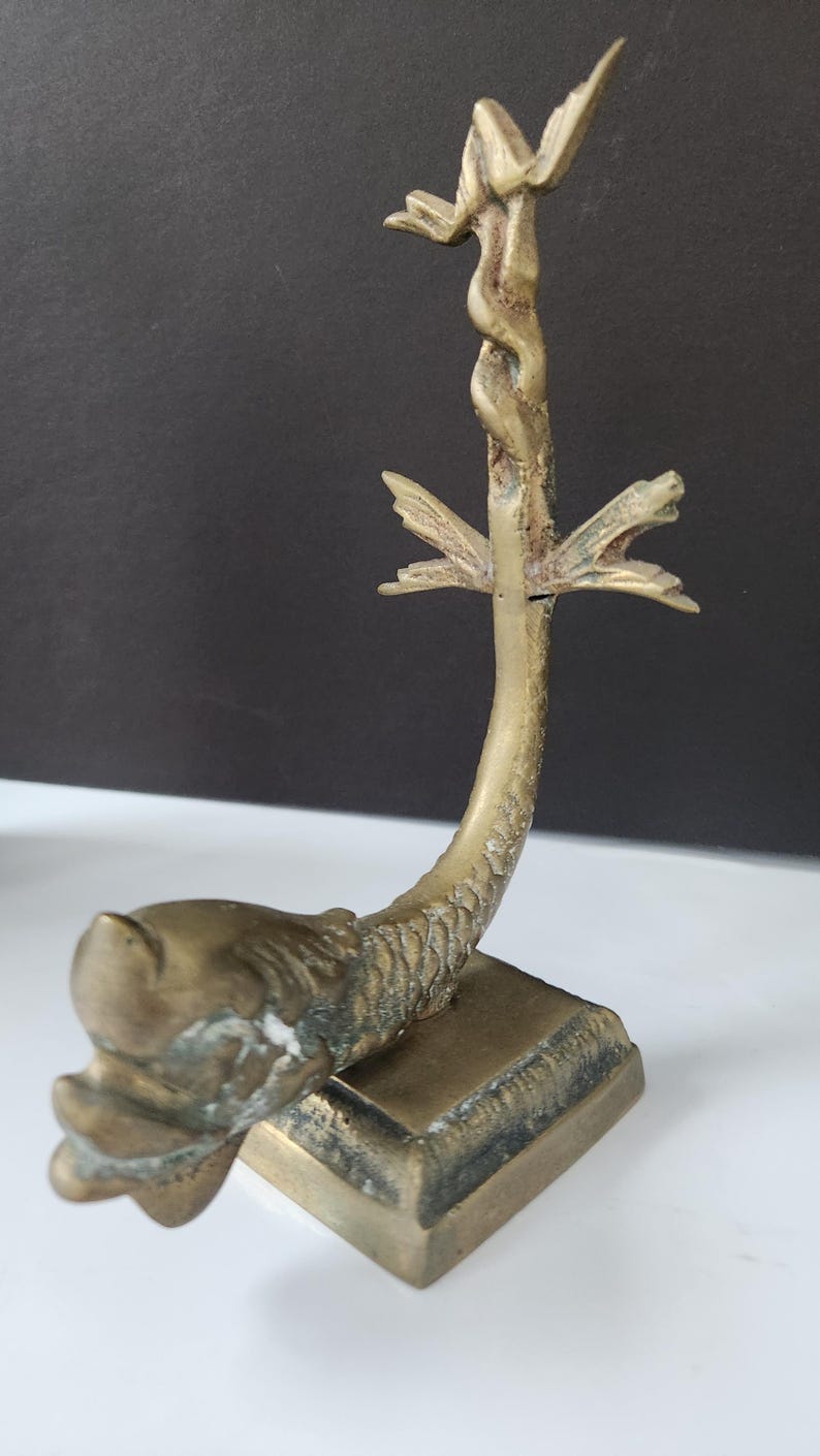 May include: A vintage brass sculpture of a mythical creature, possibly a dragon or sea serpent. The sculpture has a square base, a curved body, and a detailed head. The top features a decorative element.
