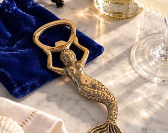May include: A gold-toned metal bottle opener shaped like a mermaid with a detailed tail and raised arms forming the opening mechanism. The bottle opener is positioned on a blue velvet drawstring pouch, showcasing a metallic finish and textured scales.