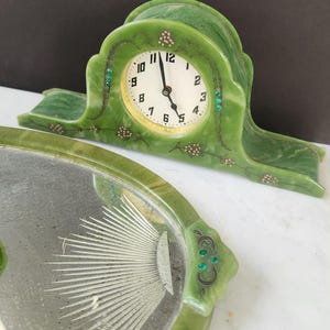 May include: A vintage green clock with a white clock face and black hands. The clock has a decorative green frame with gold and green accents. A matching green tray with a sunburst design is in the foreground.